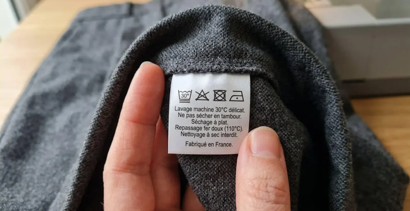 Gros plan d'une étiquette d'entretien textile affichant des symboles ISO de lavage à 30°C, interdiction sèche-linge et séchage à plat, sur un pantalon en maille gris